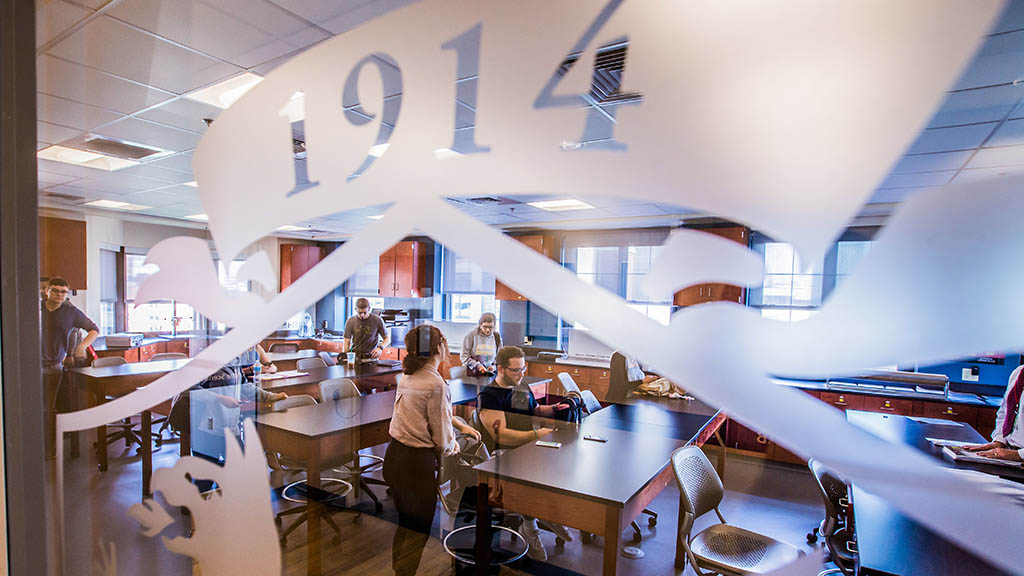 a photo taken through a classroom window of students chatting as they take seats