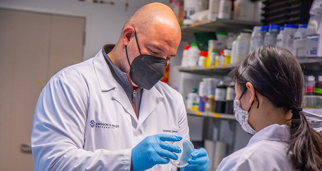 JWU Biology Professor Christos Dimos holds up a blank Petri dish while instructing a student in the research lab