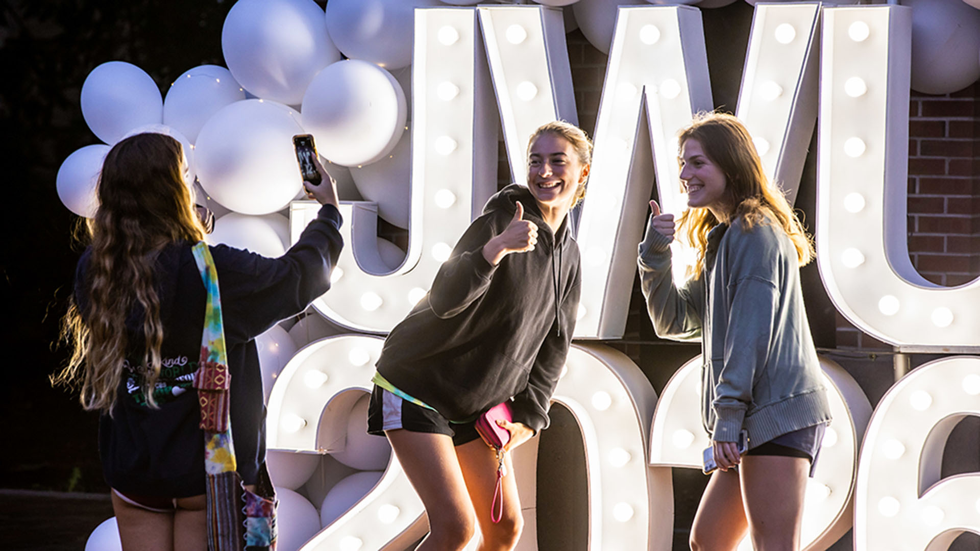 A student taking the photo of two students standing in front of a lit up JWU sign