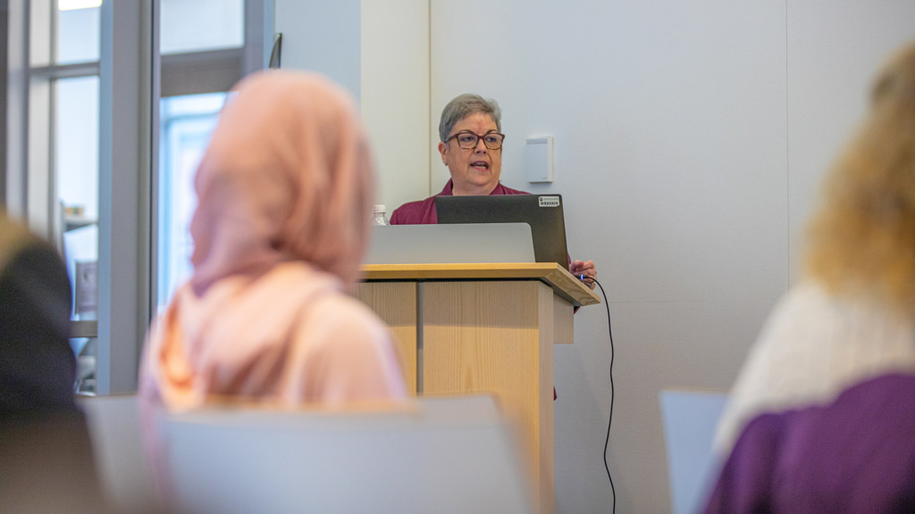 Donna Remington stands at a podium while introducing the DEIB alumni panelists