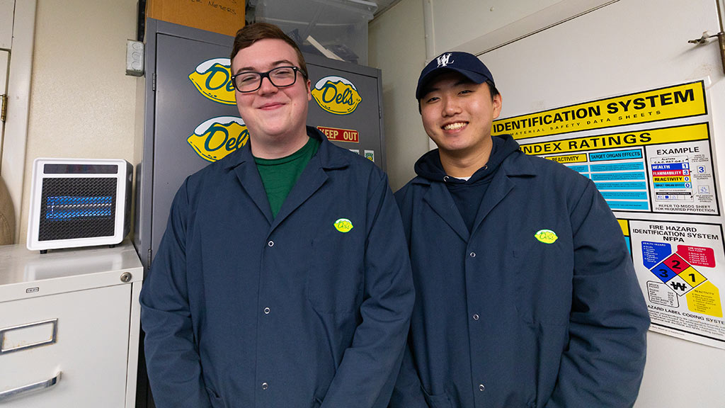 two men in matching blue lab coats pose smiling inside the lab at Del’s Lemonade