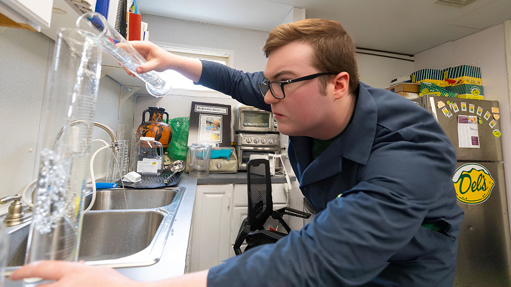 a young man in a blue lab coat concentrates as he pours liquid from one beaker into another