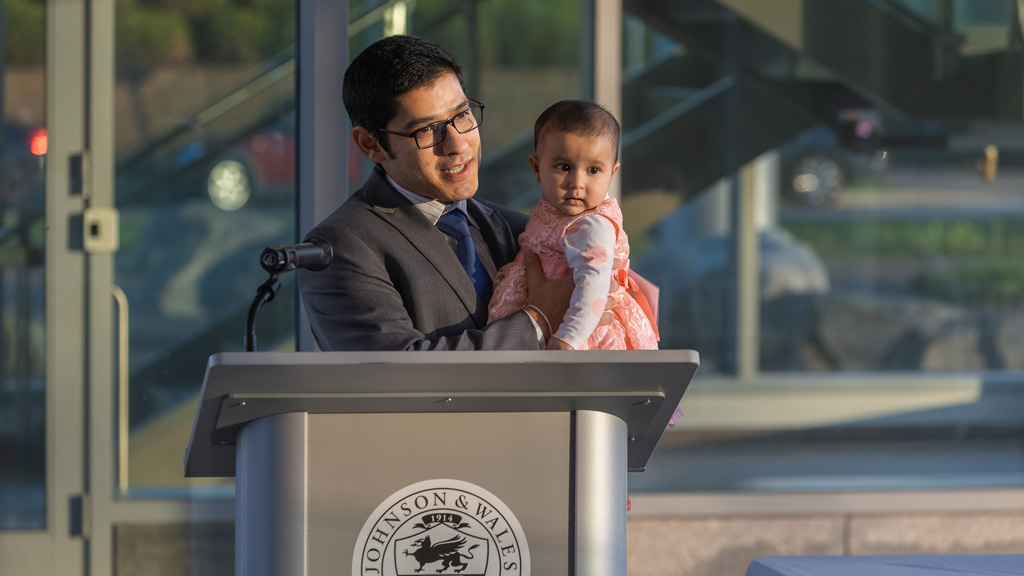 photo of Adi Dhandhania ’10 speaking at a podium while holding his daughter