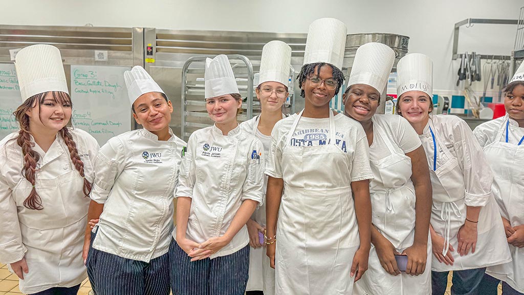 Students pose together in chef hats, white coats and aprons in a culinary kitchen lab.