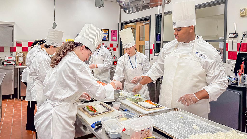 Students in white chef hats and coats layer vegetables on sushi rice to prepare sushi rolls.