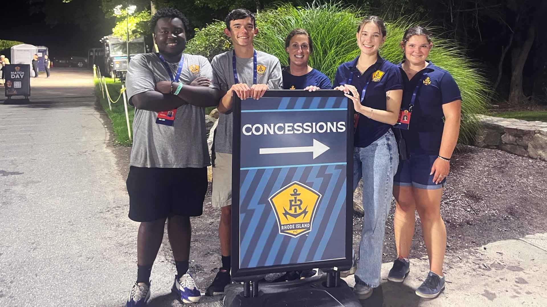 group photo taken at night of five JWU students and alum posing by a concession stand sign at a stadium