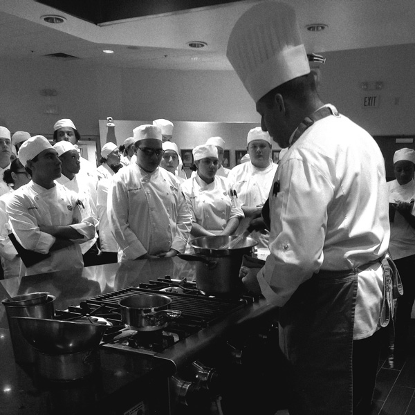 a black-and-white photo taken from behind of Dan Bazzinotti teaching a culinary class