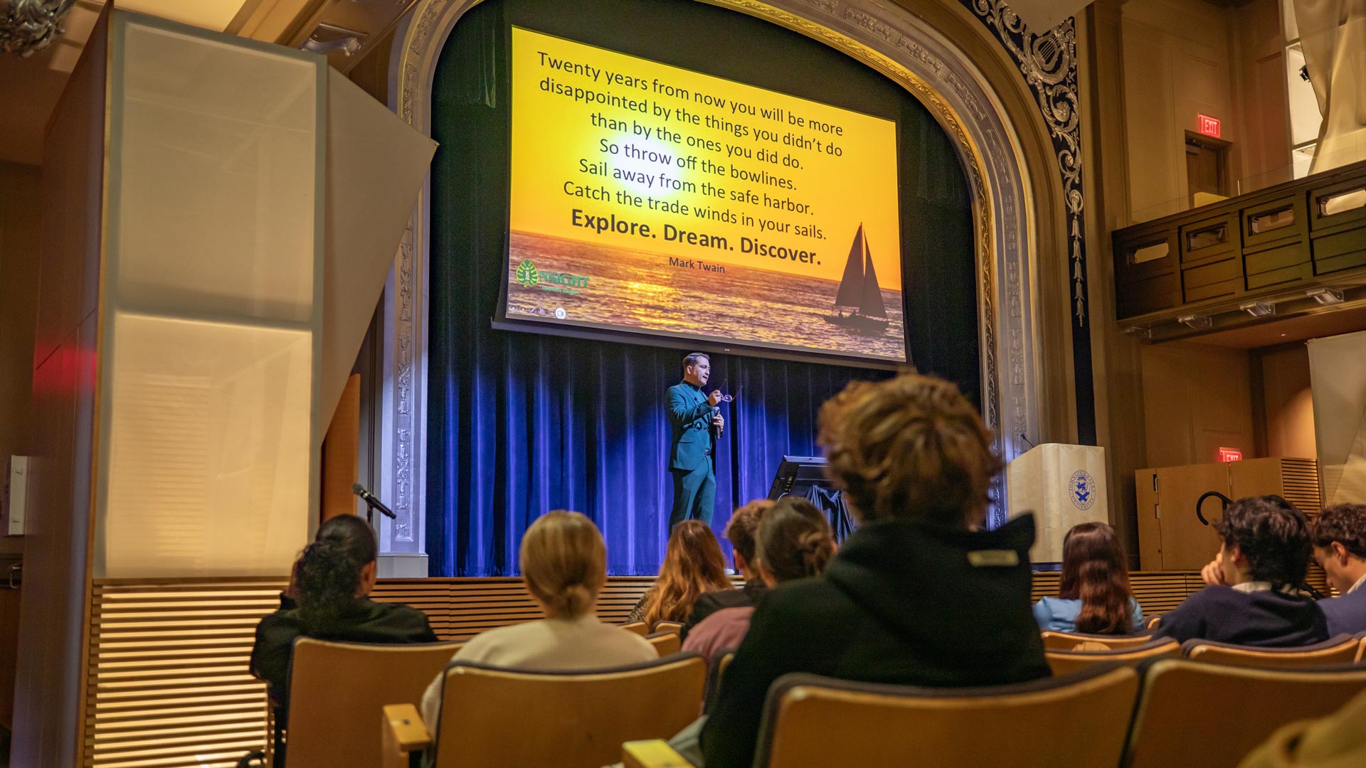 a photo showing the backs of several students' heads as they watch a speaker up on stage