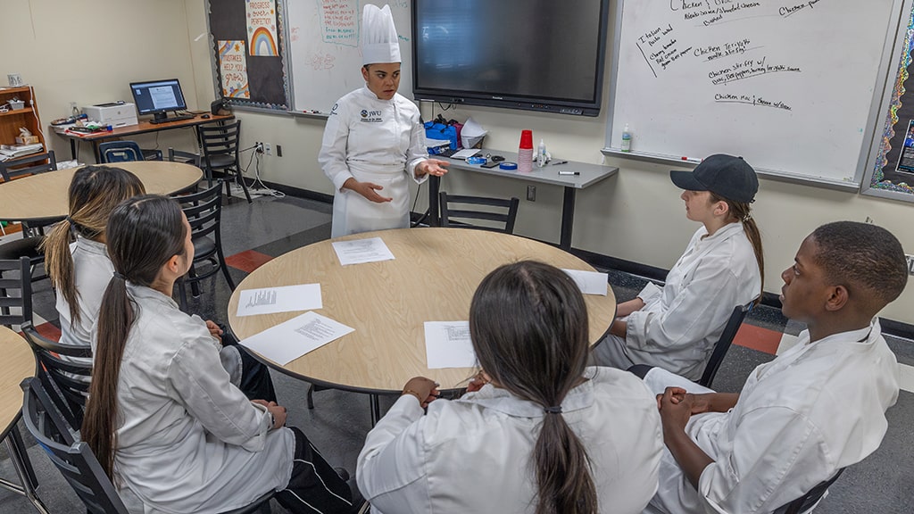 Chef instructor lectures to a group of students seated at a round table in a classroom
