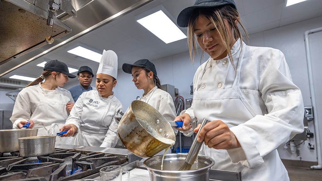 One student tempers liquids together while others watch the instructor as she helps them at the stove