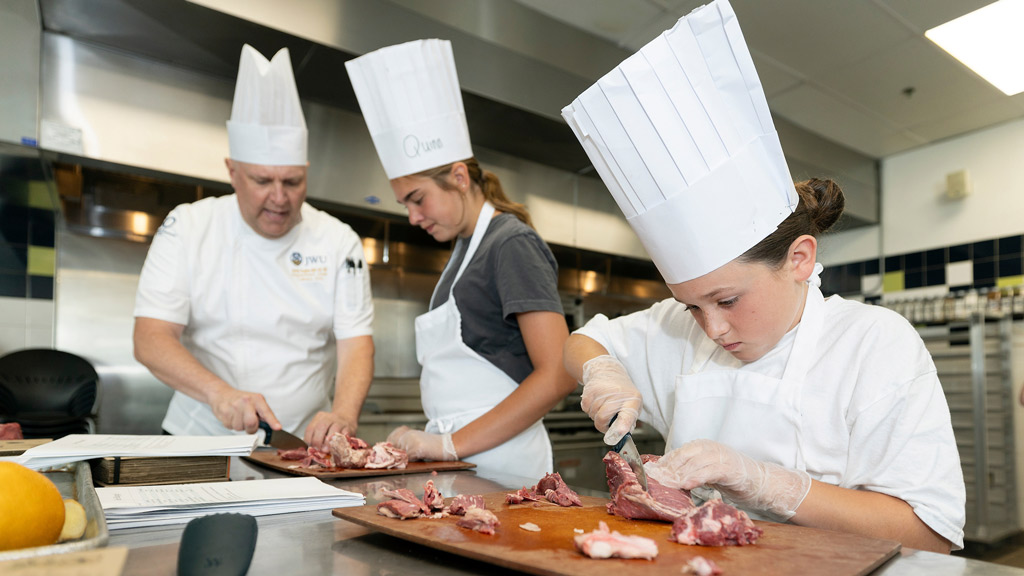 action of two girls slicing meat under the instruction of a chef