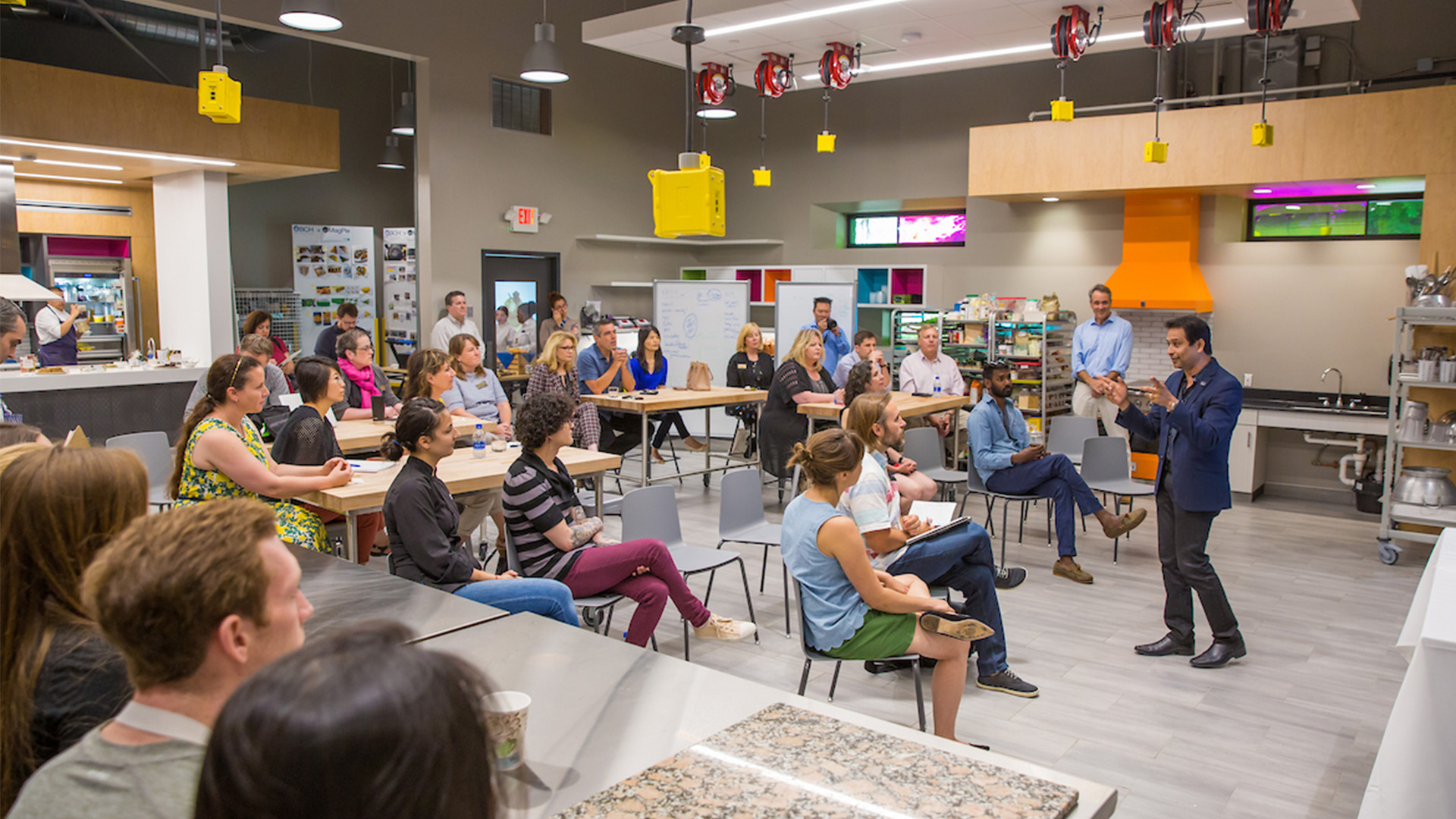 A group of attendees seated in a teaching kitchen listen to a presenter speaking at the front of the room. The space includes stainless steel counters, kitchen equipment, and a bright orange range hood.