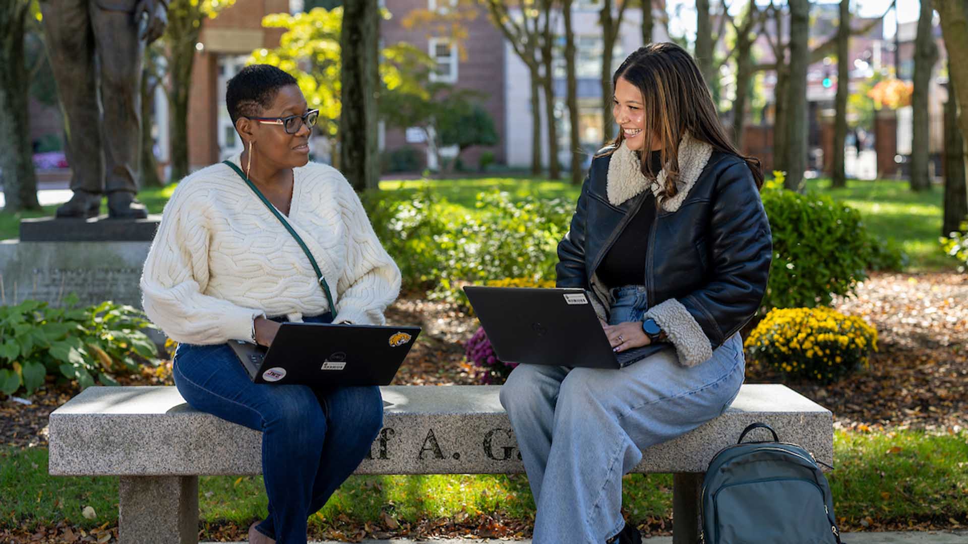 Two students sit on a bench in Gaebe Commons with their laptops.