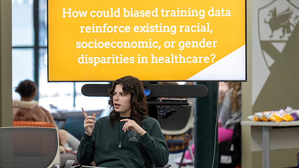 MJ Kasprzak ’26 speaks while seated in front of a PowerPoint presentation