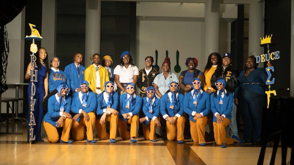 Three students, one sitting and two standing, with Greek Life shirts on smiling 